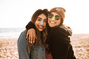 Mom with arm around daughter standing on sand with ocean in background wearing the colorful best SPF for face on their noses.